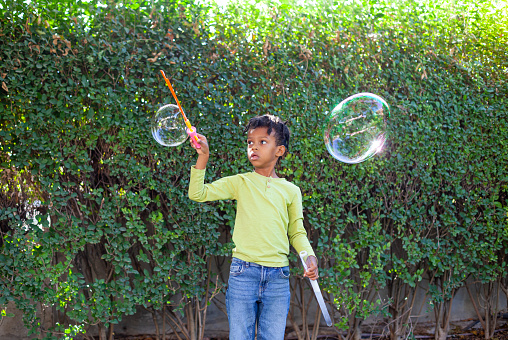 Happy African-American boy with black curly hair blowing and playing with soap bubbles at green garden