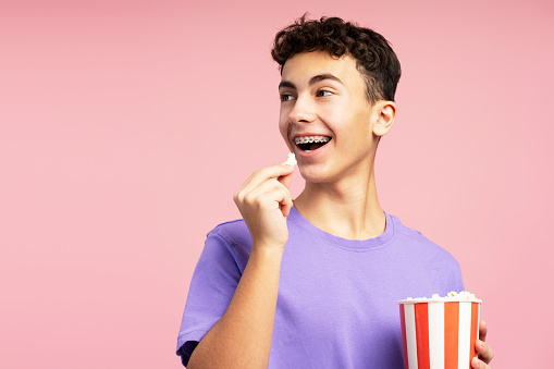 Excited, attractive teenager eating popcorn, holding bucket looking away at copy space. Handsome boy with braces watching movie. Entertainment concept