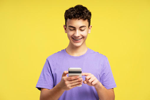 Attractive handsome teenager boy with dental braces holding mobile phone, using mobile app, ordering, text message, watching video isolated on yellow background. Technology concept, online shopping