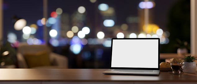 Workspace at night, a white-screen laptop computer mockup on a wooden desk in a contemporary private office room on a skyscraper. 3d render, 3d illustration
