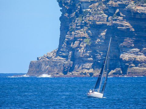 A yacht sails across Sydney Harbour with the background of the sandstone cliff face of South Head. This image was taken from Manly on a hot and sunny afternoon on 9 March 2024.