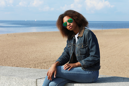 Beautiful young african woman in a denim jacket and sunglasses is resting while sitting on the beach