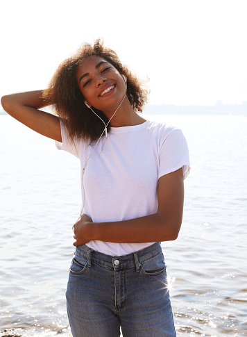 Young cute afro american woman wearing white t-shirt listening to music in headphones near sea. Summer time.