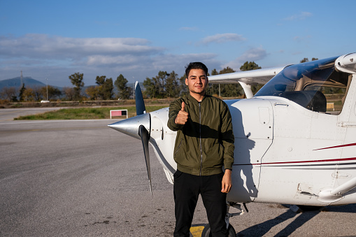 A young, handsome male pilot in casual clothes, glasses standing near a small pleasure plane on the airfield. Aviator stands on a runway near an aircraft on a landing point with private jet terminal.