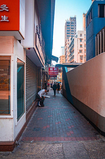 The photograph captures a moment on the bustling streets of Hong Kong, with a person bending over to arrange their belongings on the sidewalk, while pedestrians continue to navigate the narrow walkway. The closed shop shutters alongside the street and the towering buildings in the distance contrast with each other, showcasing the vibrancy of everyday life and the distinctive urban layout of the city.