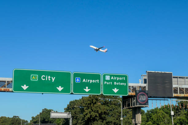 A Virgin Australia Boeing B737-8FE plane, registration VH-VUG, taking off from the third runway of Sydney Kingsford-Smith Airport as flight VA539 to the Gold Coast.  She is passing road signs on General Holmes Drive, Mascot. A surveillance camera which monitors mobile phone usage is visible above the speed limit sign. On the right are metal poles hanging down which will warn drivers of vehicles over the height limit going into the tunnel under the runway. This image was taken on a hot and sunny afternoon on 9 March 2024.