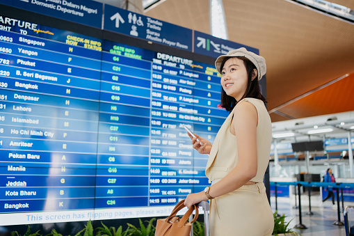Asian Chinese businesswoman with luggage using smartphone while waiting for flight at the airport. Business travel and vacation concept.