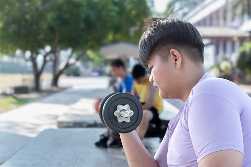 Asian chubby teenboy doing exercise with dumbbells in outdoor park in late afternoon of the day.