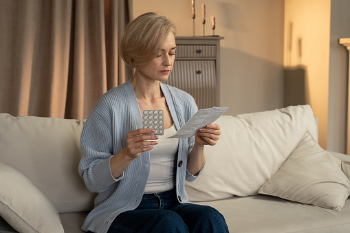 A senior woman is seated comfortably on a couch, attentively perusing a sheet of paper that appears to be associated with a blister pack of pills she is holding in her other hand, possibly checking the medication instructions.