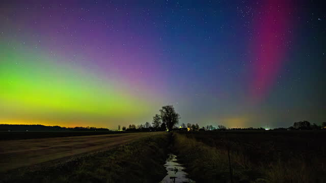 Timelapse of the vibrant Northern lights glowing above a ditch and rural fields