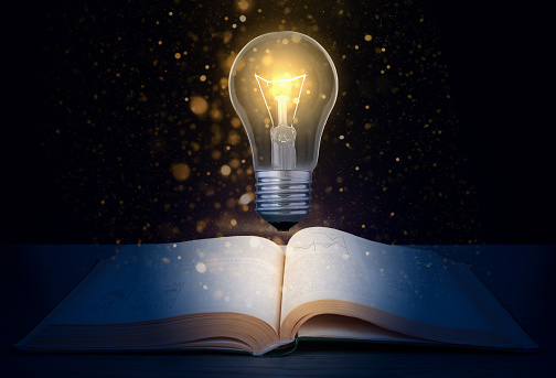 Glowing light bulb over a textbook on a dark background with glitter.  Concept to produce a new idea based on knowledge.