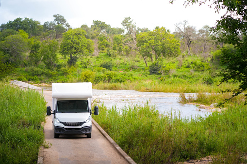 An RV faces the challenge of crossing a flooded bridge surrounded by verdant foliage. The waters of the overflowing river lap gently at the concrete structure, suggesting recent heavy rains in this serene, natural setting. The scene is peaceful yet underscores the unpredictability of rural travel.