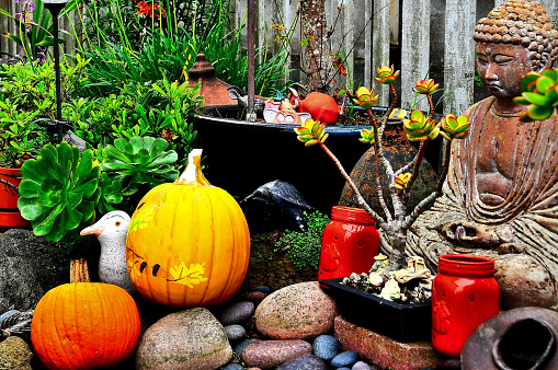 A creative image of an Oriental garden arrangement amid lush green foliage. A Buddha replica and colored stones at the foreground create a Zen-like atmosphere typical of Oriental Asian home gardens. Repurposed and recycled old materials are used to create a more distinct personal preference and identity.