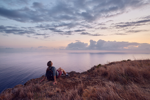 Rear view of a female backpacker day dreaming on a break from her hike while resting on cliff above the sea at sunset. Copy space.