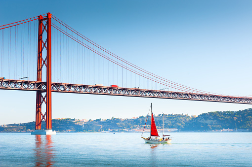 25th April Bridge over the Tejo river in Lisbon, Portugal. Yacht with red sail on the river. Famous travel destination