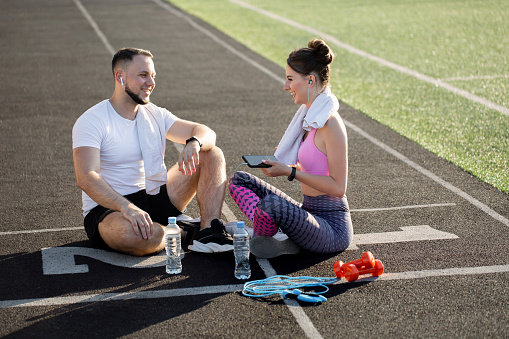 Sporty young boy and woman sit on the treadmill at the stadium in the summer after a run with a phone, headphones and sports bottles