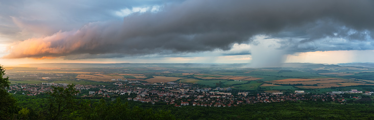 Wide angle horizontal panorama of a landscape with dramatic sky - super cell, rain storm, colored sky over a small town in a plain fieldBig rain storm is approaching small town in a plain field. Dramatic sky, rain curtain, super cell