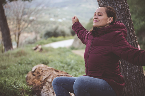 Happy young woman with closed eyes, enjoying a weekend outdoor, relaxing in nature, smiling, outstretching her arms, breathing fresh air, sitting on a log in the forest. People. Countryside lifestyle