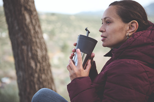Close-up side portrait of young pretty woman tourist, traveler holding stainless steel thermos mug with hot drink in the early spring forest, enjoying a happy weekend in the nature. People. Lifestyle