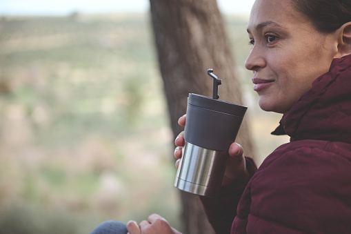 Close-up portrait of a happy smiling woman, traveler, female adventurer, tourist hiker drinking hot tea or coffee from a travel mug while resting after hiking in the nature. Copy advertising space