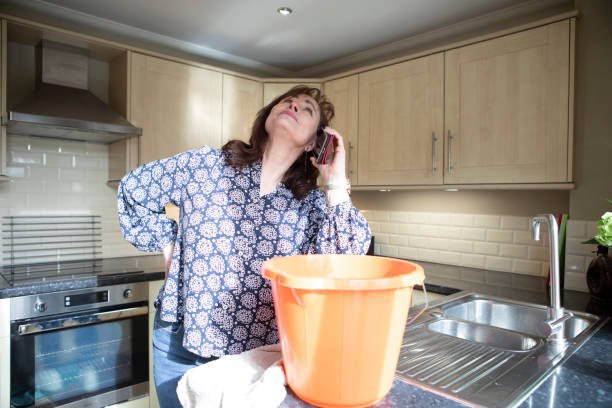 Homeowner dealing with small home insurance issues: A concerned mature female is on the telephone looking up to the ceiling with a bucket on a kitchen counter.
