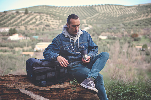 Backpacked adventurer traveler man sitting on a log in mountains, drinking his morning coffee from thermos cup, relaxing on the nature. People. Healthy lifestyle. Weekend leisure activity