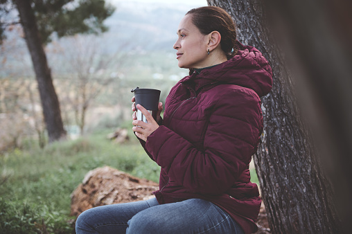 Young woman chilling out in the forest nature, holding cup of hot drink, dreamily looking into distance. People. Countryside lifestyle. Active and healthy lifestyle concept. Weekend leisure activities