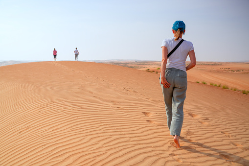 Three senior women walking, one barefoot on Wahabi desert sand, Oman. They are looking at the horizon, only sand, sky and there are some footprints in the sand.