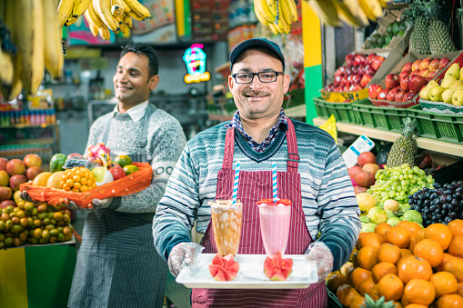 Cheerful shopkeepers sell fresh fruits, juice, smoothies, and shakes in a small store while utilizing maximum space for display. They wear hand gloves and aprons.