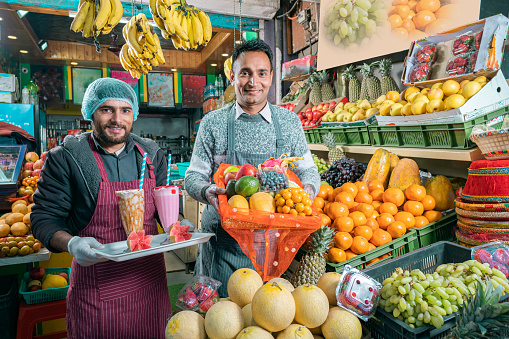 Cheerful shopkeepers sell fresh fruits, juice, smoothies, and shakes in a small store while utilizing maximum space for display. They wear hand gloves, aprons, and disposable hygiene caps.