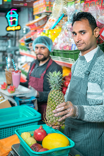 Confident shopkeepers sell fresh fruits, juice, smoothies, and shakes. They wear hand gloves and aprons.