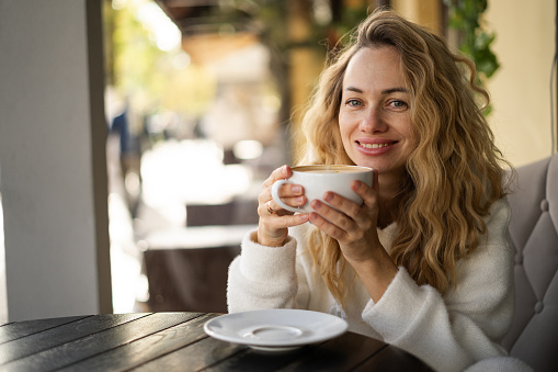 Beautiful happy blonde woman with long curly hair enjoying coffee in cafe. Copy space