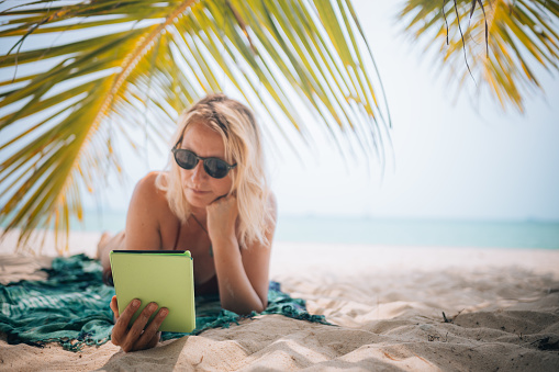 A mid adult woman relaxes under a palm tree and reads a book on an e-reader on the beach.