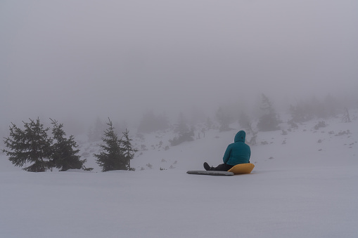 Morning from snowy mountains. A woman sits on tourist mattresses and looks at the forest. Fog. Winter