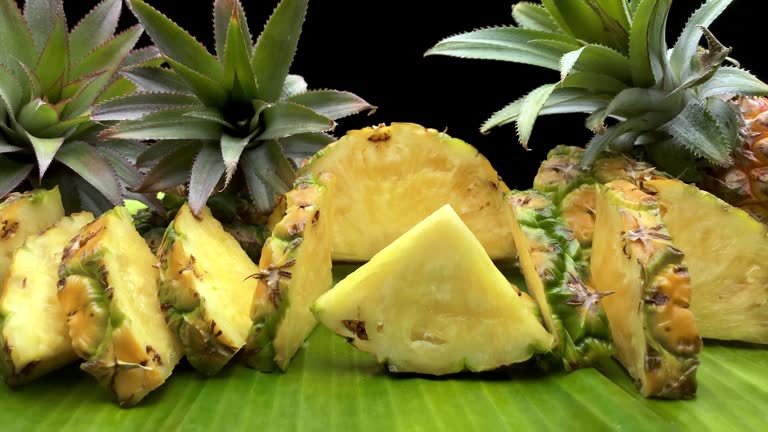 Ripe pineapple fruit slices and whole fruit on a banana leaf against a black background.