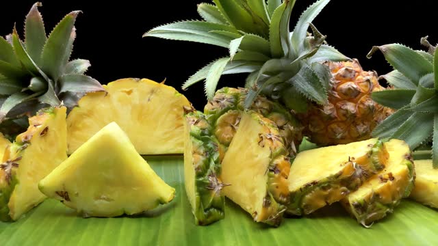 Ripe pineapple fruit slices and whole fruit on a banana leaf against a black background.