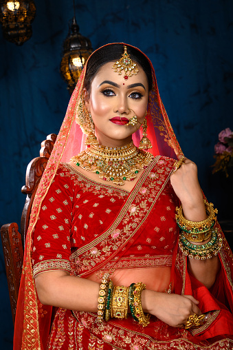 Stunning Indian bride dressed in traditional red bridal lehenga with heavy gold jewellery and veil sitting in a chair smiles tenderly in studio lighting. Wedding fashion and lifestyle.