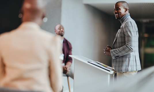 Happy business man stands confidently while presenting to colleagues in a well-lit, contemporary office space. The focus on speaker signifies leadership, success, and corporate communication.