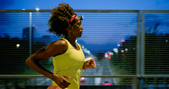 Athletic black woman running above the highway in the city at night and wearing a headlamp.
