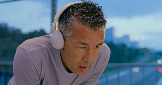 Close-up of a fit Asian man running in the city at sunset and stopping above the highway to take a breath.