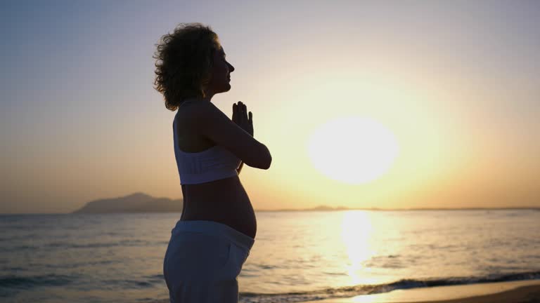 Pregnant woman is meditating at sunset on beach. Cinematic shot: atmosphere of deserted beach and pregnant woman meditating at sunset. Concept of recreation meditation at sunset on deserted beach