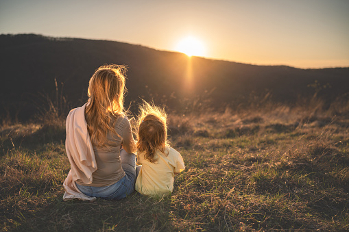 Rear view of a mother and her little daughter sit on the grass on a hill and watch the sunset