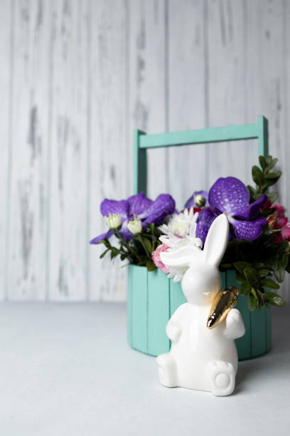 Easter bunny holding carrot, wooden basket with multicoloured flowers on concrete table, Easter background. Vertical greeting card or invitation, copy space