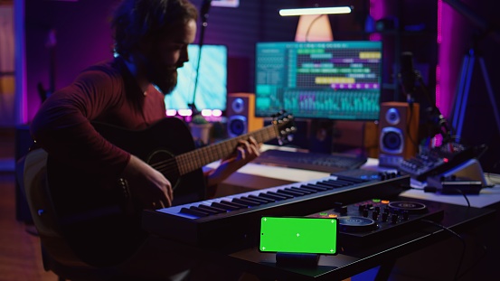Songwriter playing acoustic guitar in home studio with greenscreen display running on smartphone app. Musician practices singing on musical instrument with strings, isolated screen. Camera B.