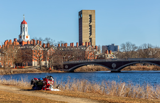 Cambridge, Massachusetts, USA - February 25, 2024: A homeless person's campsite on the Cambridge riverbank of the Charles River on a wiinter day, surrounded by their possessions on shopping carts. In the background are the student residences of Harvard University's Dunster House with its clock tower and the John Weeks bridge across the Charles River. No person is visible.