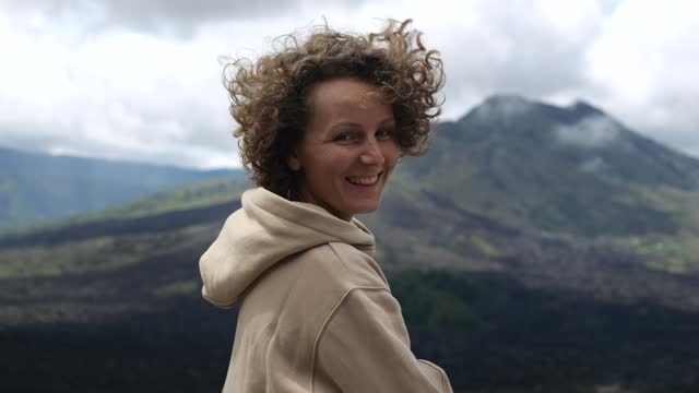 Close-up of young woman smiling to camera against beautiful mountain, curly hair blowing in wind. Captures joy of mountain travel, underlining beauty of mountain travel. Plan your mountain travel