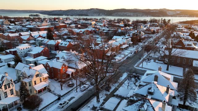Aerial view of a snow-dusted town at dusk, with a river and hills in the distance. Aerial descending shot over quaint American town after winter snow.