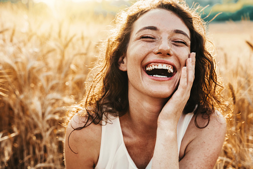 Happy beautiful woman smiling at camera in a wheat field - Delightful female enjoying summertime sunny day outside - Wellbeing, mental health, body care and happiness concept
