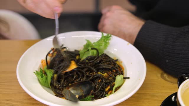 Black pasta with roasted prawns, shrimps. Served in a restaurant. Person eating pasta with fork. Black pasta and green lettuce on a white plate. Squid ink pasta with seafood and vegetables