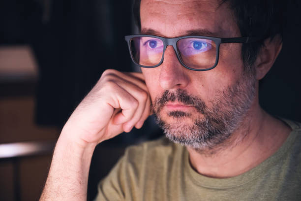 Man with eyeglasses looking at computer screen in dark room, reflection of display content on his eyewear